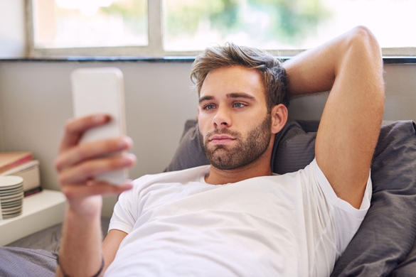Handsome young Taurus man in pajamas using a cellphone while lying in bed in the morning